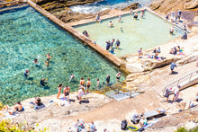 Load image into Gallery viewer, A vibrant photograph of a blue pool with clear blue water and a rocky coastline in the background, with people swimming and sunbathing.